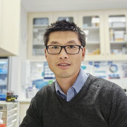 Dr Joel Zheng wears glasses and smiles at the camera with a clinical setting in the background